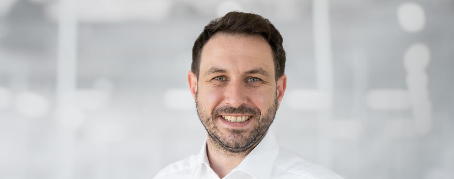 Mathias Rombach, Ausbildungsleiter lächelt in die Kamera. Eine Kollegin sitzt neben ihn und lacht.