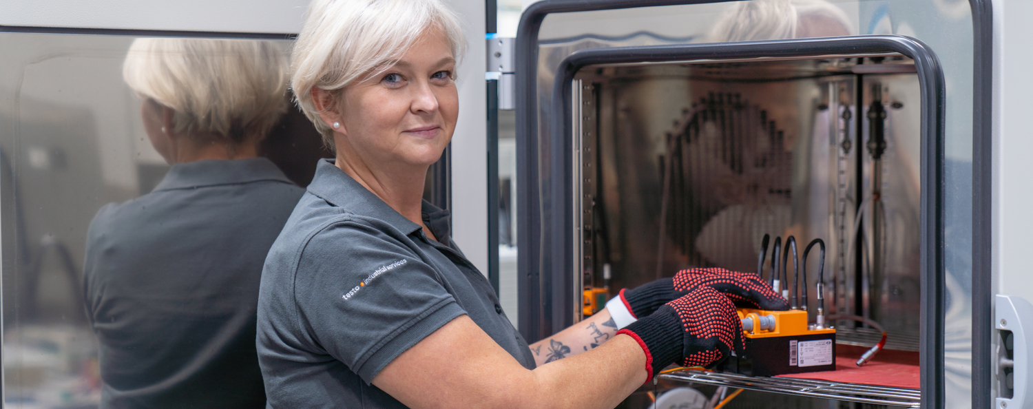 Frau mit blonder Kurzhaarfrisur steht halb seitlich zur Kamera. Nebenher hantiert Sie an einem Klimaschrank.