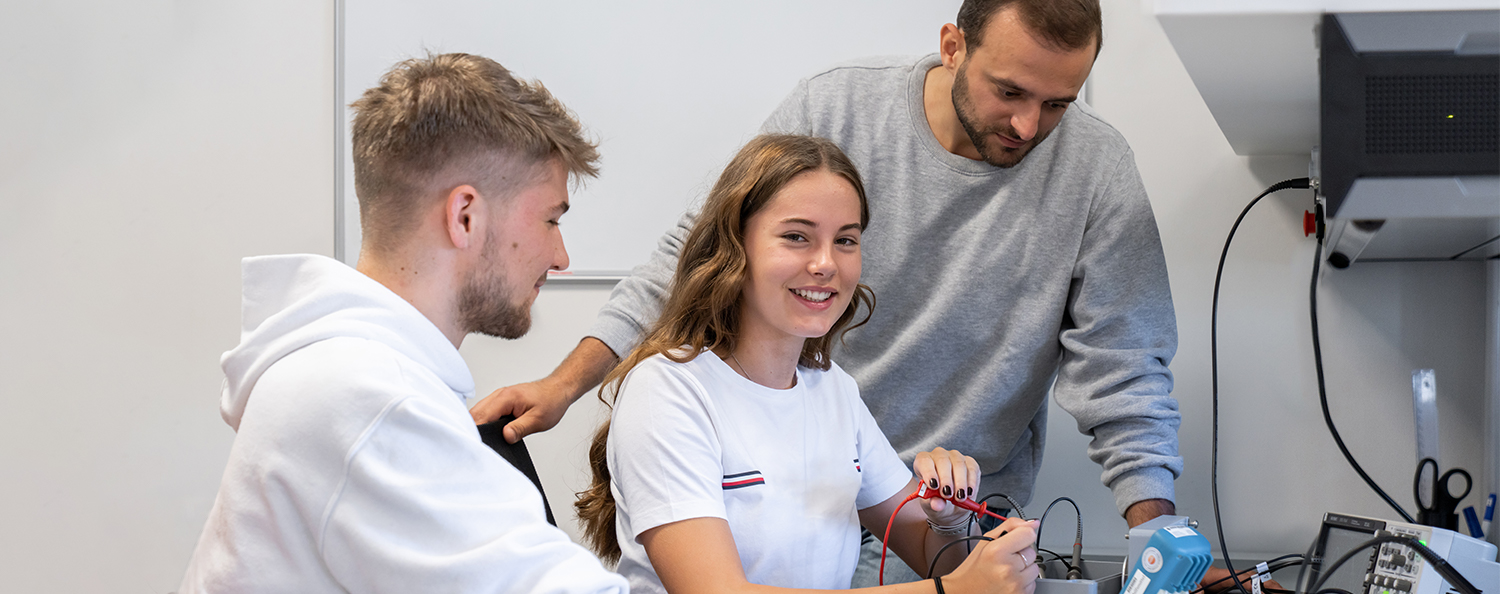 Ein Mitarbeiter erklärt zwei Praktikanten einen technischen Laboraufbau.