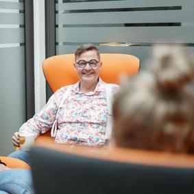 Person sitzt in einem modernen Bürobereich auf einem orangefarbenen Sessel und hält ein Getränk, Gesprächssituation in entspannter Umgebung