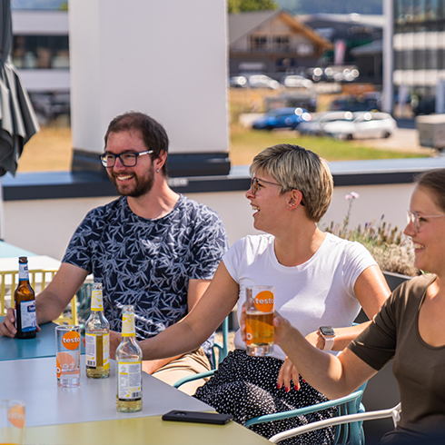 Mike, Jacqueline & Monika sitzen am Tisch, lachen in die Richtung ihrer Freunde und trinken ein Radler.