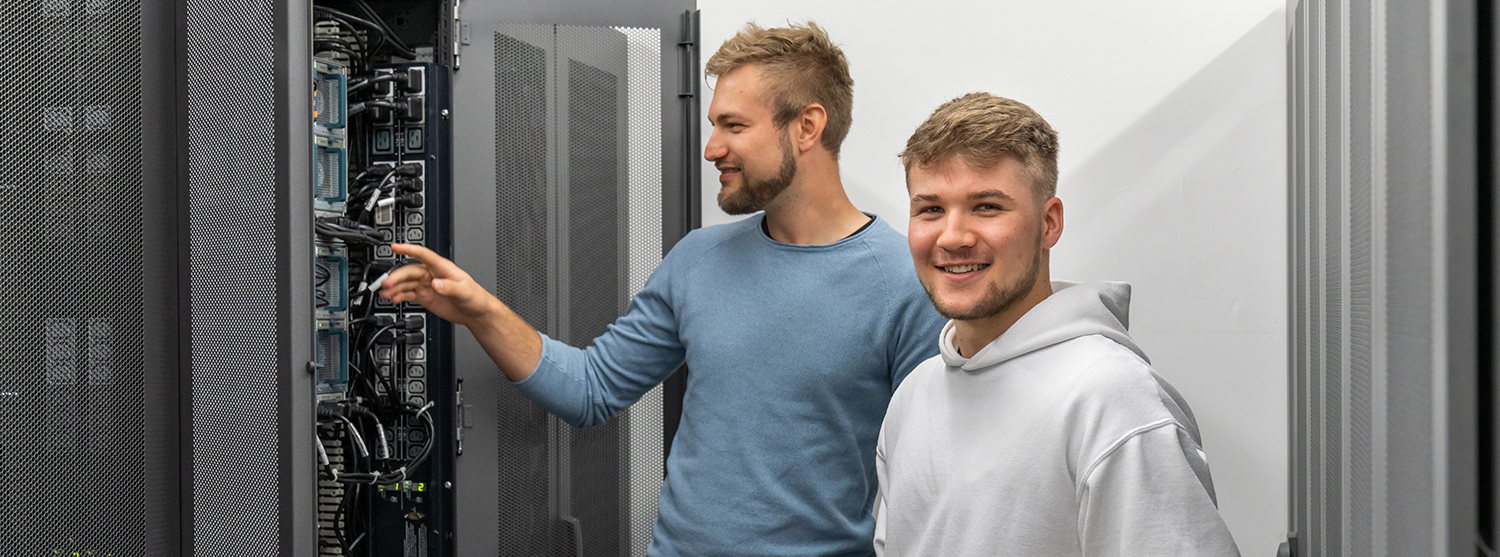 Ein angehender Fachinformatiker steht an einem Serverschrank und lässt sich von seinem Ausbilder etwas erklären. 