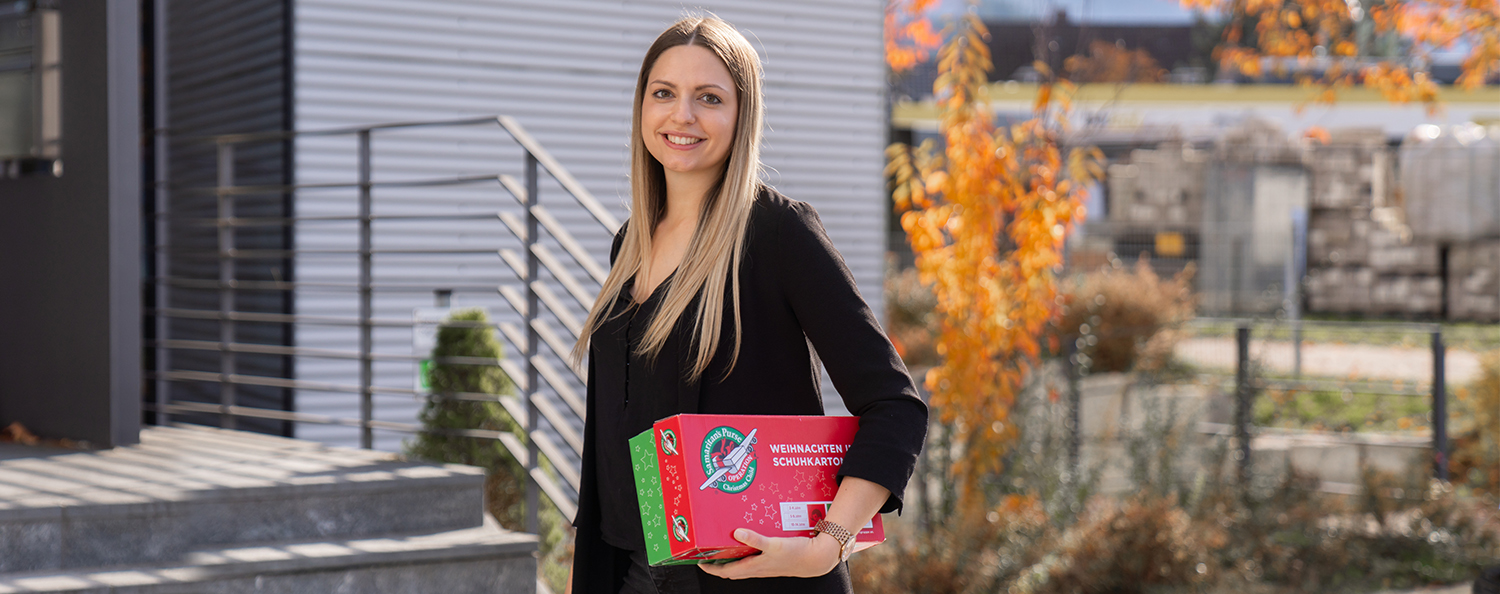 Eine Mitarbeiterin hält einen offiziellen Karton in der Hand der Geschenkeaktion "Weihnachten im Schuhkarton" und lächelt in die Kamera.