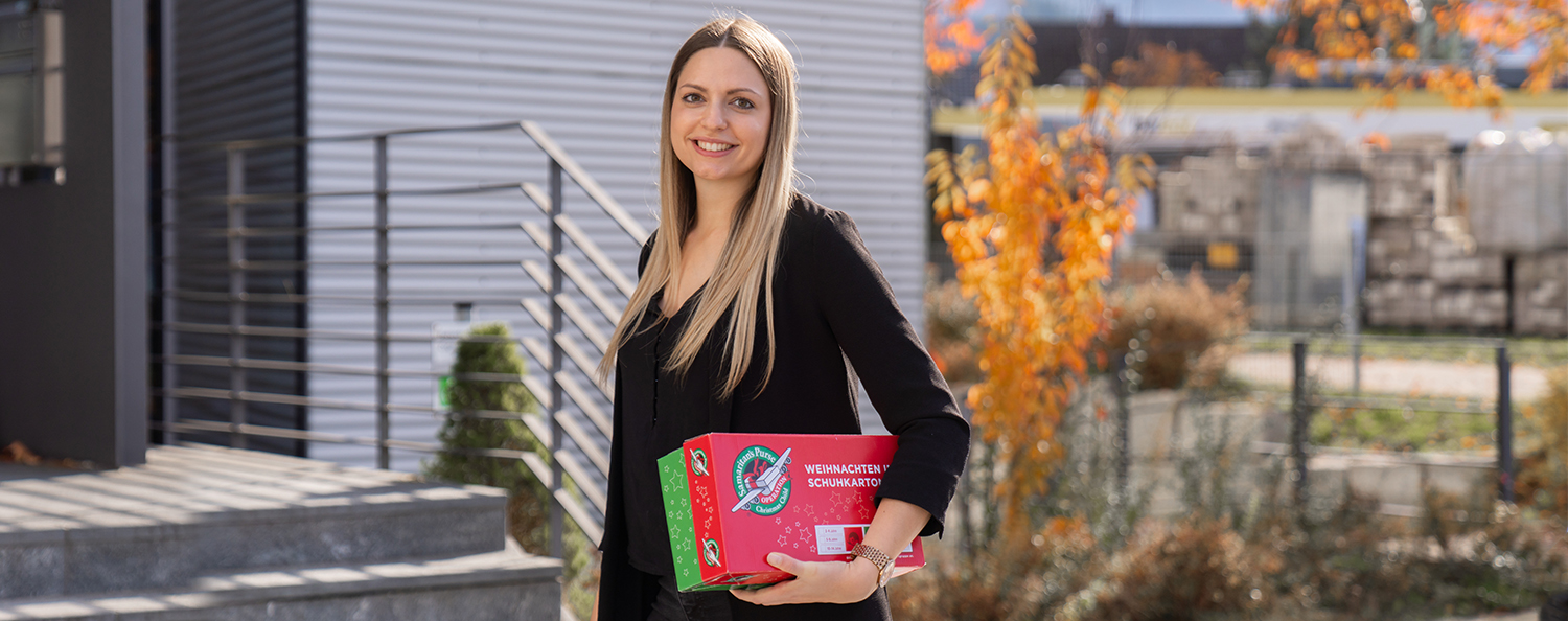 Geschenkeaktion "Weihnachten im Schuhkarton" Eine Mitarbeiterin hält einen offiziellen Karton in der Hand der Geschenkeaktion "Weihnachten im Schuhkarton" und lächelt in die Kamera.