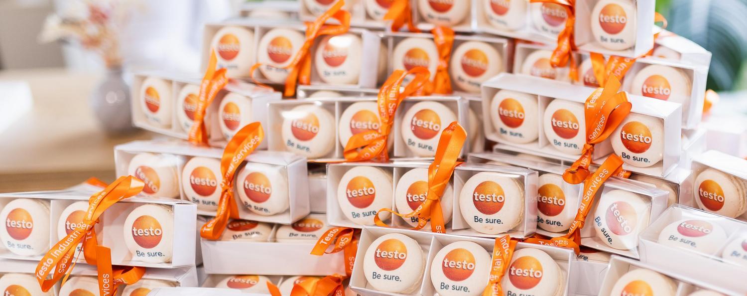 Symposium Kalibrierung & Prüfmittelmanagement 2026 Auf einem Tisch liegen zahlreiche kleine Geschenkboxen mit Macarons mit Testo-Logo und orangefarbenen Schleifen.