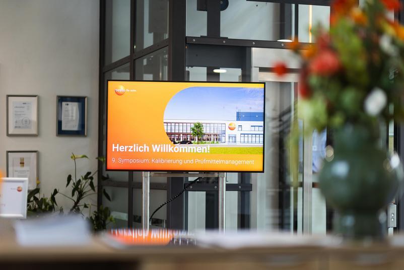 Symposium Kalibrierung & Prüfmittelmanagement 2026 Ein Bildschirm im Eingangsbereich einer Veranstaltung zeigt die Begrüßungsfolie zum Symposium Kalibrierung und Prüfmittelmanagement. Im Hintergrund sind Glaswände und Pflanzen zu sehen.