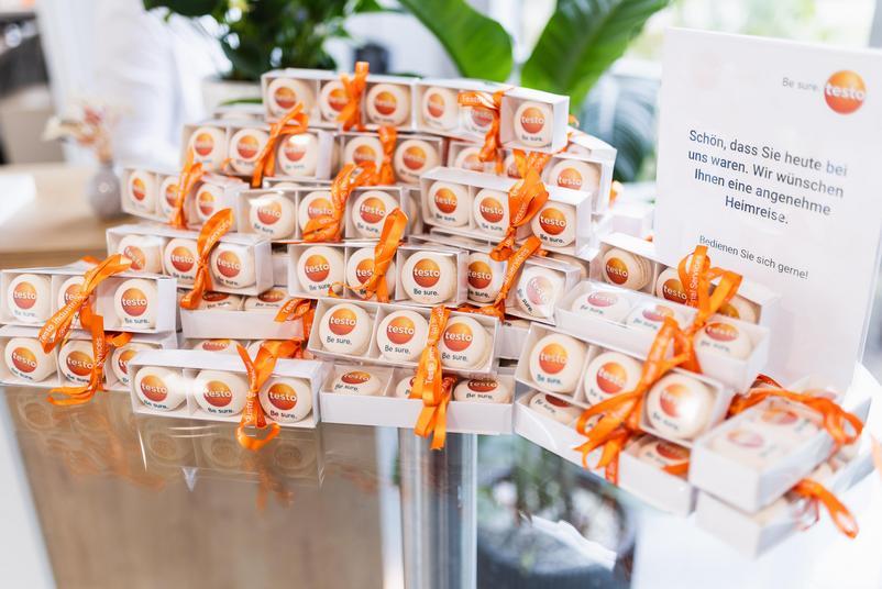 Symposium Kalibrierung & Prüfmittelmanagement 2026 Auf einem Tisch liegen zahlreiche kleine Geschenkboxen mit Macarons mit Testo-Logo und orangefarbenen Schleifen. Daneben informiert ein Aufsteller die Teilnehmenden über ein Abschiedsgeschenk zur Mitnahme.