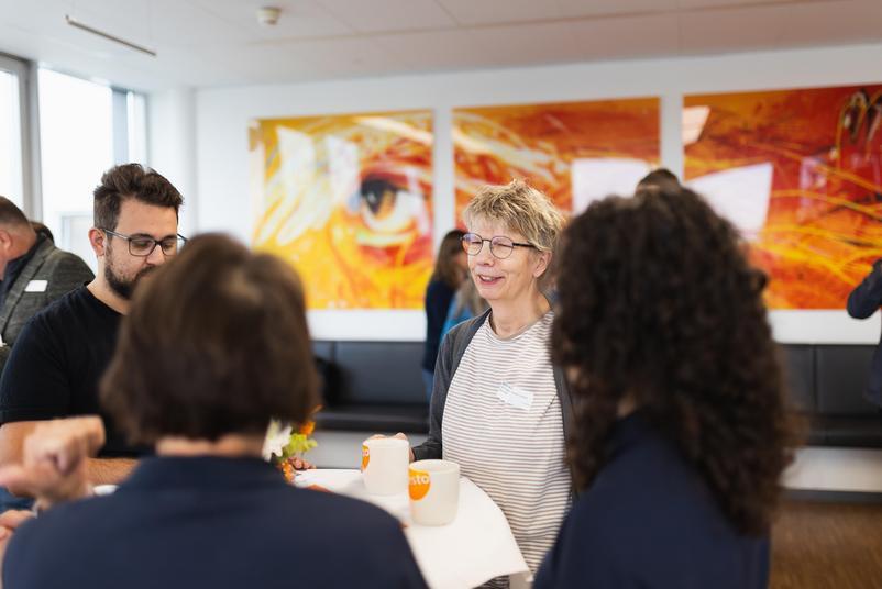 Symposium Kalibrierung & Prüfmittelmanagement 2026 Mehrere Teilnehmende sitzen und stehen an einem Stehtisch im Gespräch. Im Hintergrund hängen großformatige Wandbilder in warmen Orange- und Gelbtönen.