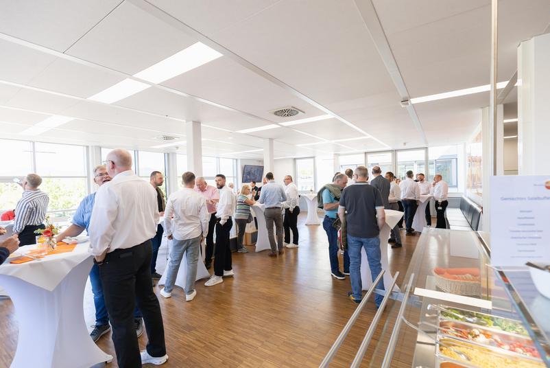 Symposium Kalibrierung & Prüfmittelmanagement 2026 Mehrere Teilnehmende stehen in einem hellen Veranstaltungsraum mit großen Fenstern und Holzboden an Stehtischen zusammen und führen Gespräche. Im Vordergrund ist ein Buffetbereich mit Salaten zu sehen.