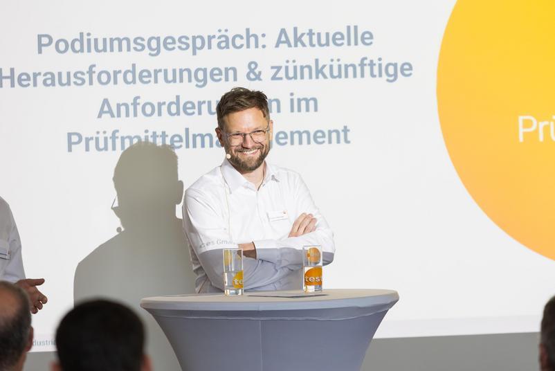 Symposium Kalibrierung & Prüfmittelmanagement 2026 Eine Person steht während eines Podiumsgesprächs hinter einem Stehtisch auf einer Bühne. Im Hintergrund ist eine Präsentationsfolie zum Thema Herausforderungen und zukünftige Anforderungen im Prüfmittelmanagement zu sehen.