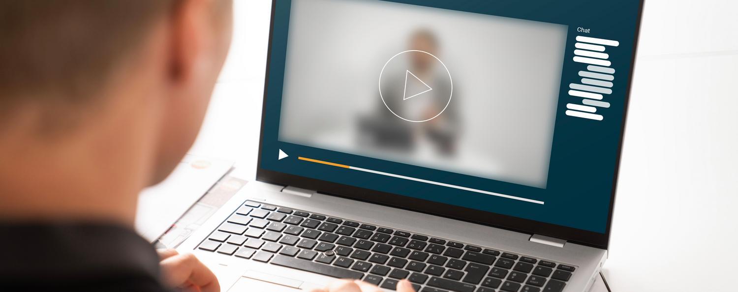Rückansicht eines Mannes der auf den Laptop vor sich schaut. Auf dem Laptop sieht man eine unscharfe Person in einer Webinarsituation.