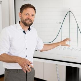 Mann mit Hemd steht vor einer Leinwand, auf derer eine Kurve einer Statistik zu sehen ist.