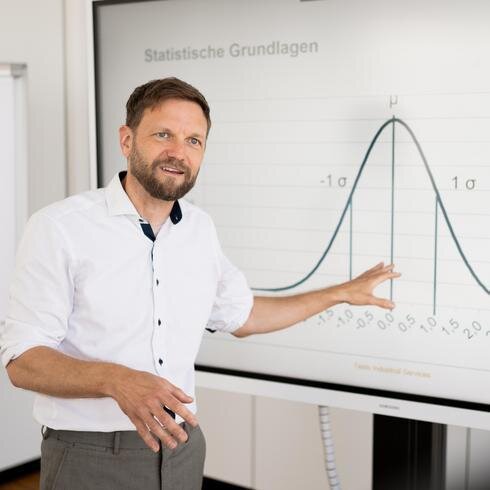 Mann mit Hemd steht vor einer Leinwand, auf derer eine Kurve einer Statistik zu sehen ist.